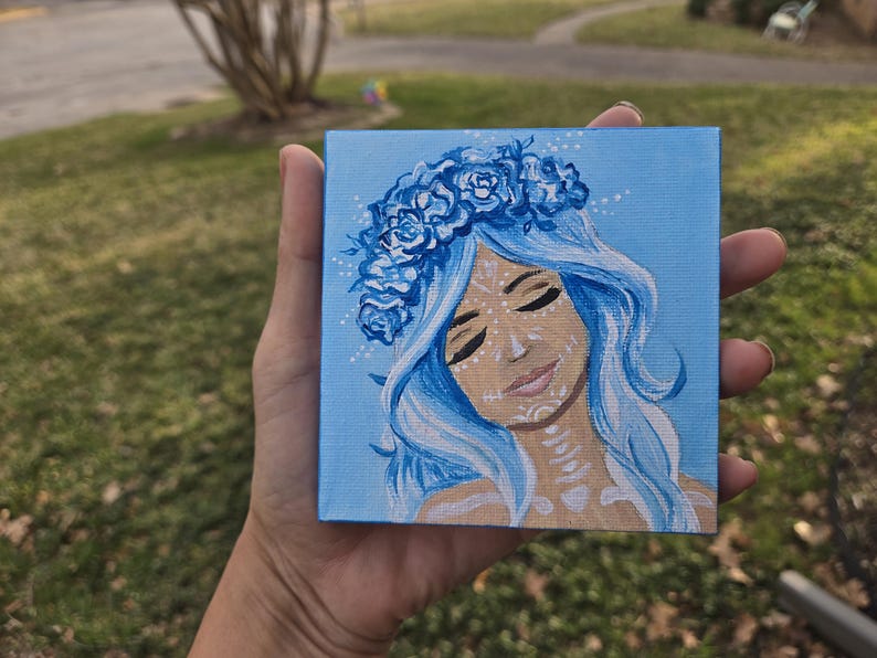 May include: A small square canvas painting depicting a woman with blue hair and a floral crown. The artwork has a light blue background and the woman's face has white decorative markings. The painting is held in a hand.
