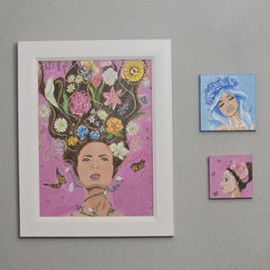 May include: A framed artwork showing a woman with flowers in her hair, butterflies, and a pink background. Two smaller square paintings depict women with floral accents. The art pieces are displayed on a grey wall.