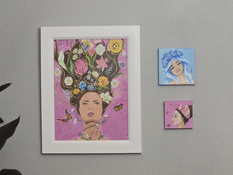 May include: A framed artwork showing a woman with flowers in her hair, butterflies, and a pink background. Two smaller square paintings depict women with floral accents. The art pieces are displayed on a grey wall.