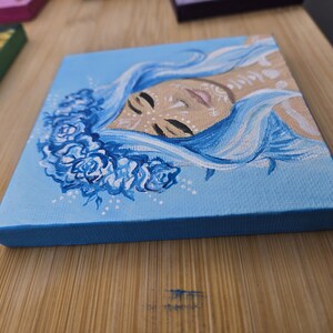 May include: Small square canvas art depicting a face with flowing blue hair and a floral crown. The painting uses shades of blue, beige, and white. The canvas has a light blue surface and a dark blue edge.