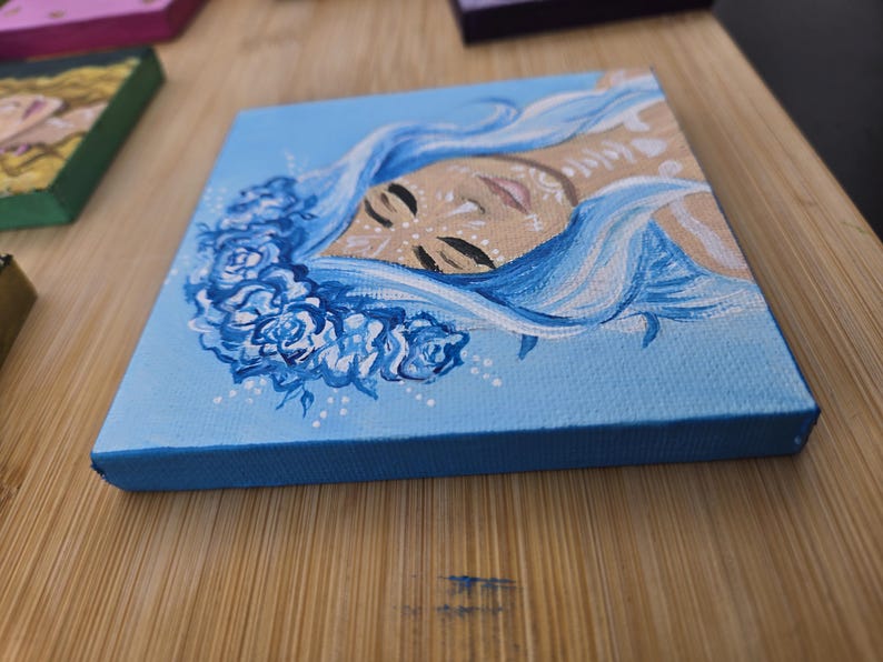 May include: Small square canvas art depicting a face with flowing blue hair and a floral crown. The painting uses shades of blue, beige, and white. The canvas has a light blue surface and a dark blue edge.