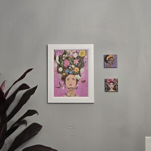 May include: Three framed artworks on a grey wall. The largest is a white-framed print of a woman with flowers in her hair. Two smaller square canvases depict women with floral headwear. A houseplant is in the lower left.