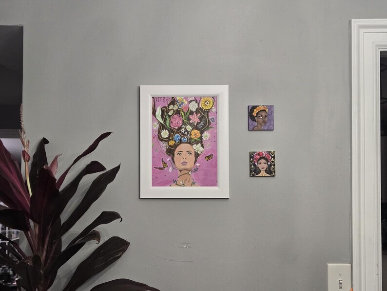 May include: Three framed artworks on a grey wall. The largest is a white-framed print of a woman with flowers in her hair. Two smaller square canvases depict women with floral headwear. A houseplant is in the lower left.