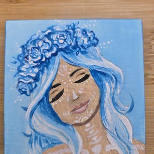 May include: Square canvas art depicting a woman with blue hair and a floral crown. The artwork shows a face with closed eyes and white decorative markings. The background is a light blue, and the style is artistic and decorative.