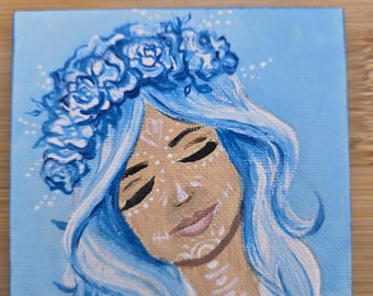May include: Square canvas art featuring a serene woman with blue hair and a floral crown. The artwork depicts a face with closed eyes and white decorative markings. The background is a light blue, and the overall style is artistic and decorative.