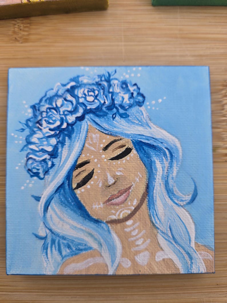 May include: Square canvas art depicting a woman with blue hair and a floral crown. The artwork shows a face with closed eyes and white decorative markings. The background is a light blue, and the style is artistic and decorative.