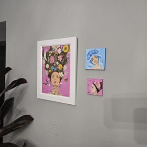 May include: Three artworks are displayed on a grey wall. The largest is a framed painting of a woman adorned with flowers. Two smaller square paintings show women with floral headpieces, one blue and one pink.