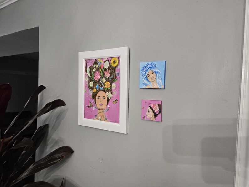 May include: Three artworks are displayed on a grey wall. The largest is a framed painting of a woman adorned with flowers. Two smaller square paintings show women with floral headpieces, one blue and one pink.