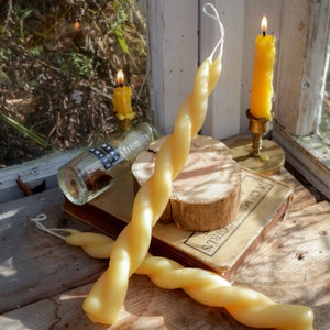 May include: Two pale yellow beeswax taper candles with a twisted spiral design. One candle stands upright against a wooden slice, while the other lies horizontally across an old book. A small glass bottle labeled STAR ANISE sits nearby.