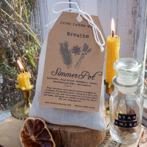 Peut inclure: Un sac en mousseline blanche rempli d'un mélange d'herbes et d'épices pour un pot mijoté. Le sac est muni d'une étiquette en papier brun avec le texte "crow takes wing Breathe Simmer Pot Lavender, Star Anise, Rosemary, Lemon, Marshmallow Root, Astragalus Clove Pour contents into medium sized saucepan or crockpot filled with water. Bring to a boil and simmer on low. Let the aromatics fill your heart and home. Add water as needed to enjoy for hours. Not for consumption. www.crowtakeawing.com @crow.takes.wing". Il y a un petit bocal en verre contenant de l'anis étoilé et deux bougies jaunes.