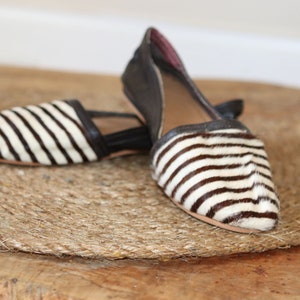 Op de afbeelding: Een paar zwarte en witte gestreepte leren platte schoenen met een spitse neus. De schoenen liggen op een geweven kleed.