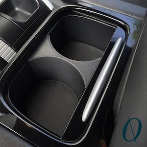 Puede incluir: Un portavasos de consola de coche negro con un acento plateado. El portavasos tiene dos compartimentos circulares y un compartimento rectangular. El acento plateado recorre el lateral del portavasos.