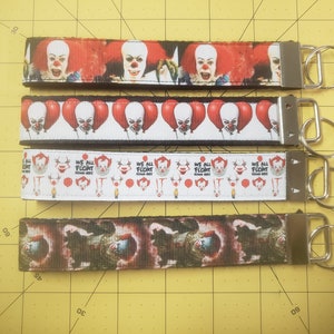 May include: Four keychains with metal hardware. Each keychain features a ribbon with a horror-themed design. The designs include images of a clown with red hair and balloons, and the text "WE ALL FLOAT DOWN HERE".
