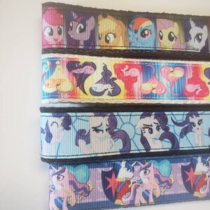 May include: Ribbon featuring colorful cartoon character designs. The ribbon has a black border and is stacked in rows. The characters are in various colors, including pink, purple, yellow, blue, and white. The ribbon is likely used for crafting or decorating.