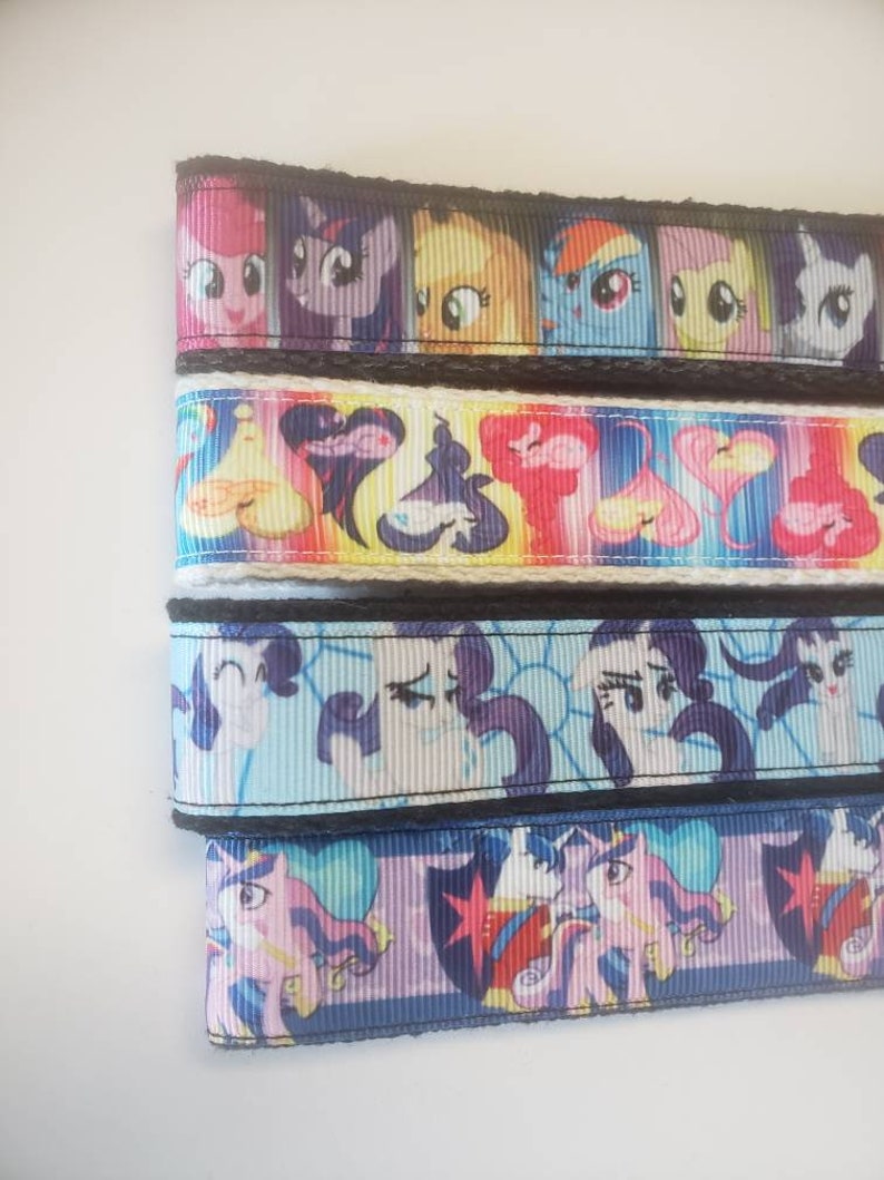 May include: Ribbon featuring colorful cartoon character designs. The ribbon has a black border and is stacked in rows. The characters are in various colors, including pink, purple, yellow, blue, and white. The ribbon is likely used for crafting or decorating.