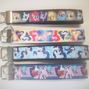 May include: Four keychains with colorful cartoon character designs. Each keychain features a fabric strap with a metal key ring. The designs include various characters in bright colors, set against black, white, and blue backgrounds.