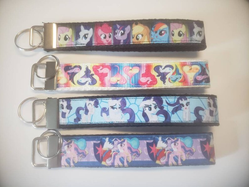 May include: Four keychains with colorful cartoon character designs. Each keychain features a fabric strap with a metal key ring. The designs include various characters in bright colors, set against black, white, and blue backgrounds.