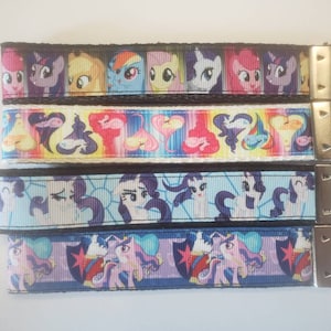 May include: Four keychains with colorful cartoon pony designs. Each keychain has a different pattern, including images of the ponies and the word "PLAY". The keychains have silver metal hardware.