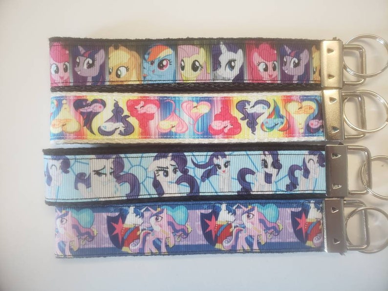 May include: Four keychains with colorful cartoon pony designs. Each keychain has a different pattern, including images of the ponies and the word "PLAY". The keychains have silver metal hardware.