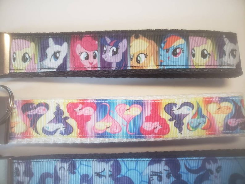 May include: Three colorful keychains featuring cartoon pony characters. The top keychain has a black border and a series of pony faces. The middle keychain has a white border with heart and pony designs. The bottom keychain has a blue border with pony faces.