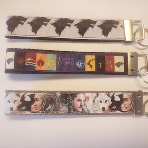 Puede incluir: Tres llaveros con herrajes plateados. El llavero superior tiene un fondo blanco con cabezas de lobo negras. El llavero del medio tiene las palabras "Game of Thrones" y varios escudos de casas. El llavero inferior tiene imágenes de personajes.