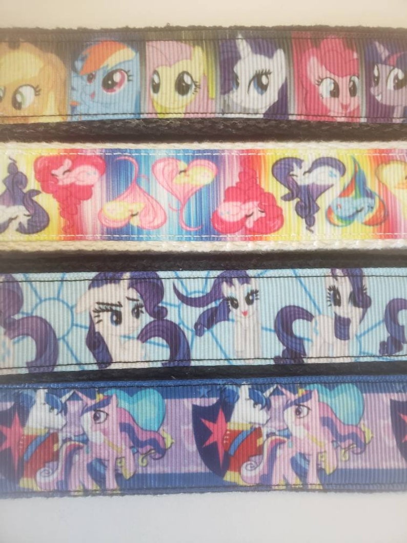 May include: Close-up of several ribbons featuring colorful cartoon pony characters. The ribbons display various designs, including pony faces, hearts, and other graphics. The ribbons are bordered with black lace.