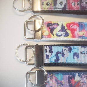 May include: Four keychains with colorful cartoon character designs. Each keychain has a silver-colored metal clasp and ring. The designs feature various characters on a black, white, and blue background. The keychains are likely made of a durable fabric.