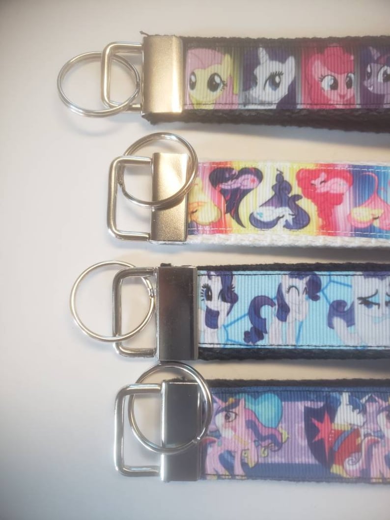 May include: Four keychains with colorful cartoon character designs. Each keychain has a silver-colored metal clasp and ring. The designs feature various characters on a black, white, and blue background. The keychains are likely made of a durable fabric.