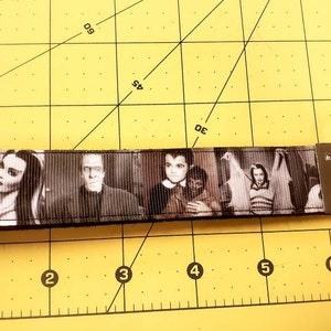 May include: Black and white key fob featuring images of characters from the classic television show, The Munsters. The key fob has a metal clasp and a silver key ring. The background is a yellow cutting mat.