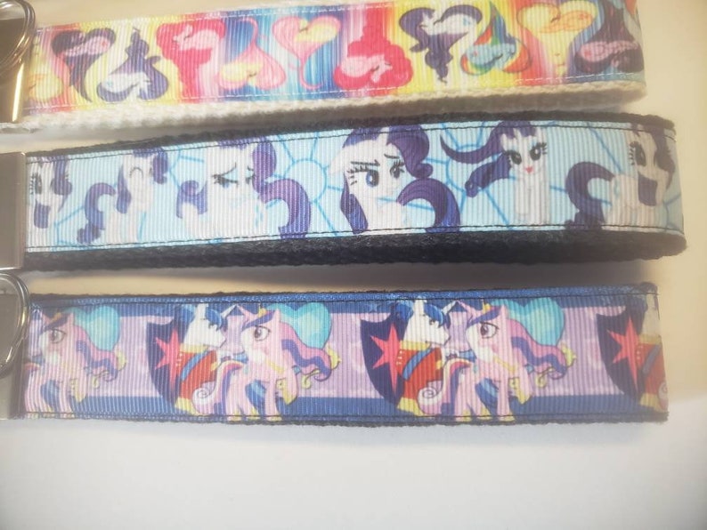 May include: Three keychains with colorful cartoon pony designs. The top keychain features a rainbow pattern, the middle one has a light blue background, and the bottom one has a blue background. Each keychain has a metal clasp.