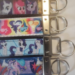 May include: Four keychains with colorful cartoon pony designs. Each keychain features a different pattern with images of the ponies. The keychains have a silver metal clasp and a key ring.