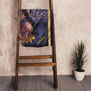May include: A decorative blanket with a fantasy art design, featuring a figure with a long braid, set against a blue and gold background. The blanket is draped over a wooden ladder, with a plant in a white pot to the side.