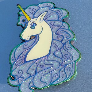 Puede incluir: Un unicornio blanco con un cuerno amarillo y una melena azul con purpurina iridiscente. El unicornio est&aacute; mirando hacia la izquierda y tiene una expresi&oacute;n caprichosa.