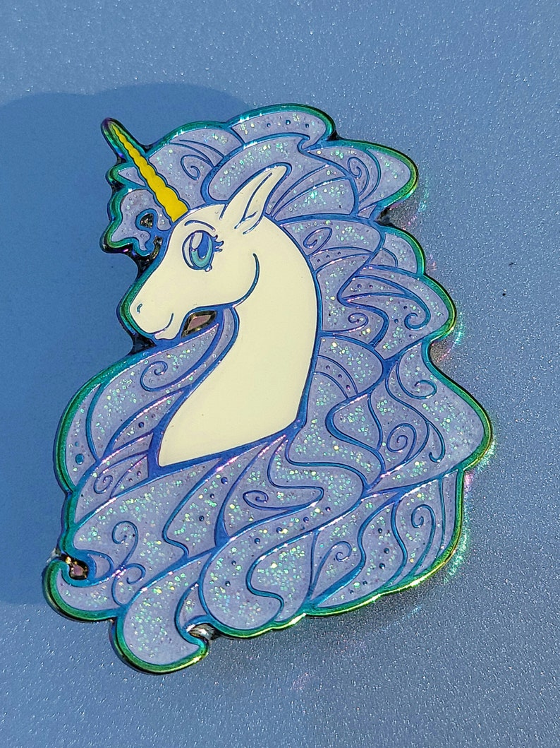 Puede incluir: Un unicornio blanco con un cuerno amarillo y una melena azul con purpurina iridiscente. El unicornio est&aacute; mirando hacia la izquierda y tiene una expresi&oacute;n caprichosa.