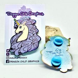 Puede incluir: Un pin de esmalte morado y blanco con un unicornio con una melena fluida y un solo cuerno. El pin tiene un acabado brillante y est&aacute; respaldado con un cierre de goma azul.