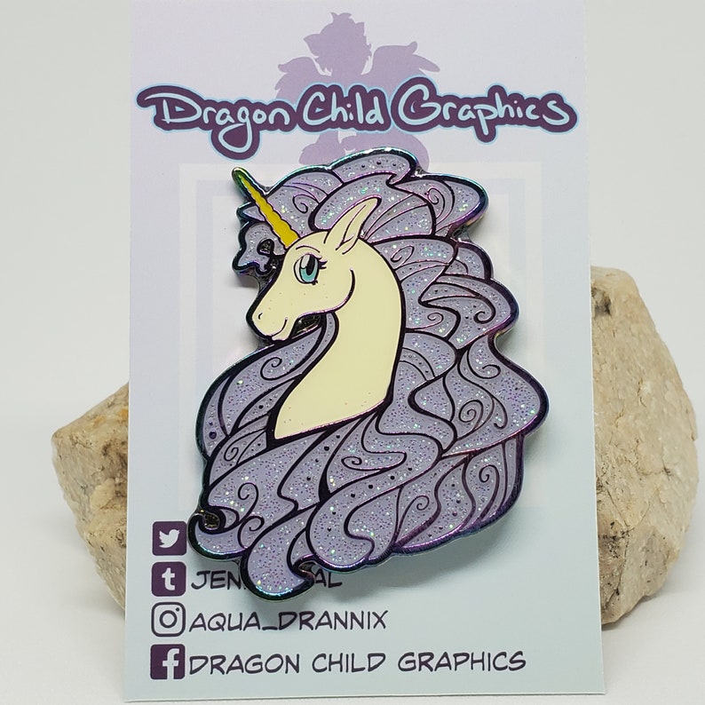 Puede incluir: Pin de esmalte de purpurina morado con un unicornio blanco con un cuerno amarillo y ojos azules. La crin y la cola del unicornio son fluidas y est&aacute;n detalladas con remolinos. El pin est&aacute; en una tarjeta blanca con el texto "Dragon Child Graphics" en negro.