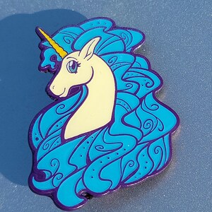 Puede incluir: Un pin de esmalte azul y morado con un unicornio blanco con un cuerno amarillo y una crin azul. La crin del unicornio est&aacute; dise&ntilde;ada en un patr&oacute;n de remolino.