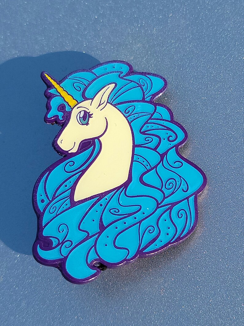 Puede incluir: Un pin de esmalte azul y morado con un unicornio blanco con un cuerno amarillo y una crin azul. La crin del unicornio est&aacute; dise&ntilde;ada en un patr&oacute;n de remolino.