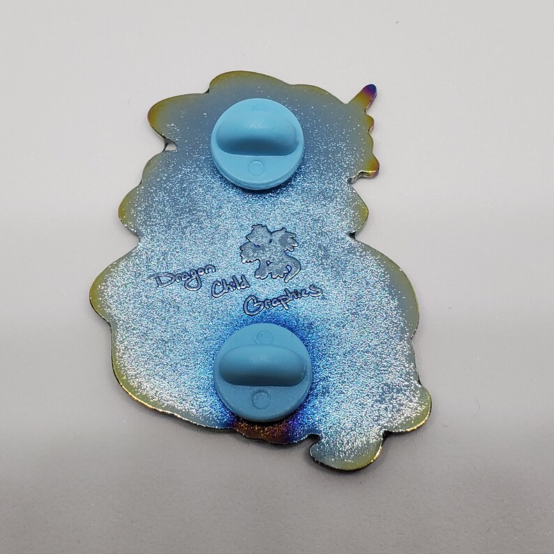 Puede incluir: Un pin de esmalte azul e iridiscente con un drag&oacute;n con una corona y el texto "Dragon Child Graphics" en la parte posterior.