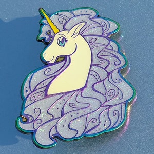 Puede incluir: Un unicornio blanco con un cuerno dorado y una melena y cola iridiscentes de color p&uacute;rpura. El unicornio mira hacia la izquierda y tiene un ojo azul. La melena y la cola est&aacute;n detalladas con remolinos y purpurina.