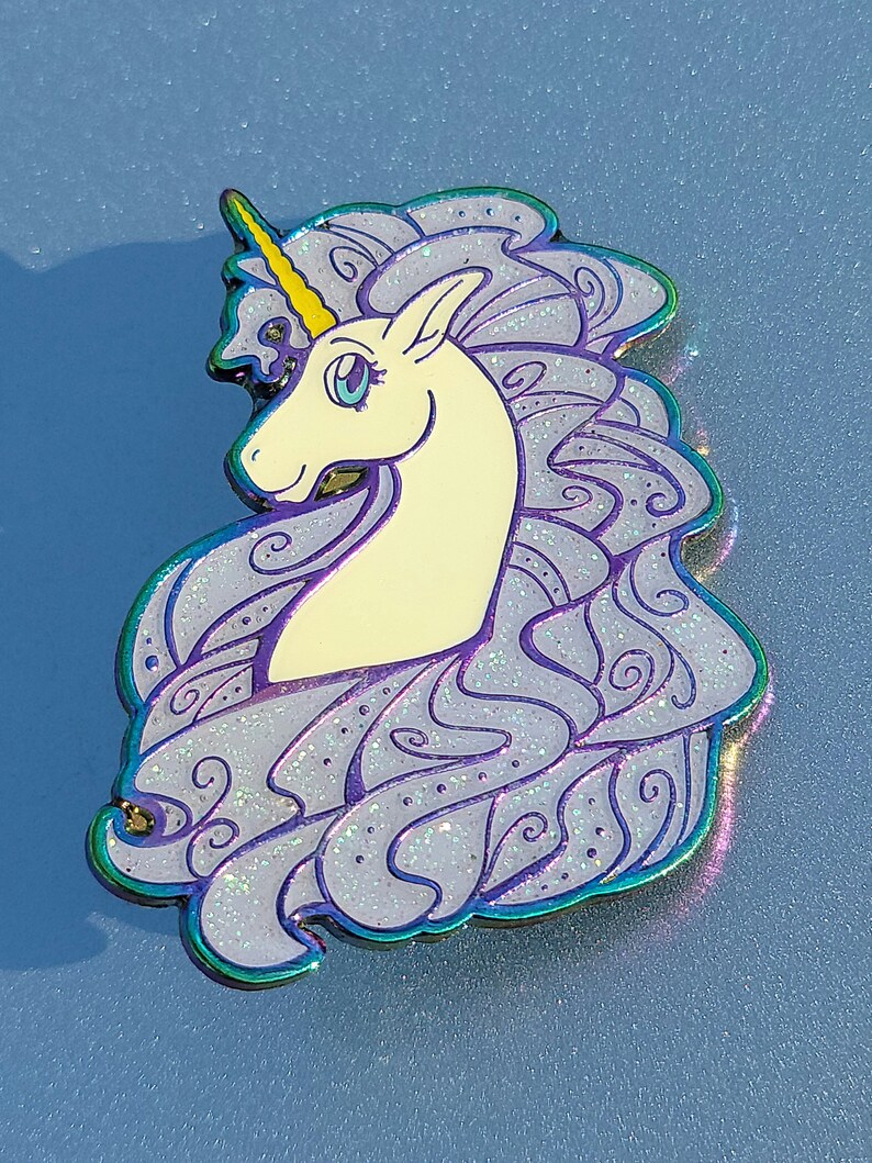 Puede incluir: Un unicornio blanco con un cuerno dorado y una melena y cola iridiscentes de color p&uacute;rpura. El unicornio mira hacia la izquierda y tiene un ojo azul. La melena y la cola est&aacute;n detalladas con remolinos y purpurina.