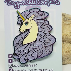 Puede incluir: Un pin de esmalte morado y blanco con un unicornio con un cuerno dorado y una crin brillante. El pin es una vista de perfil de la cabeza y el cuello del unicornio. El fondo es una tarjeta blanca con el texto "Dragon Child Graphics" en negro.