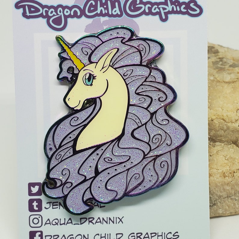 Puede incluir: Un pin de esmalte morado y blanco con un unicornio con un cuerno dorado y una crin brillante. El pin es una vista de perfil de la cabeza y el cuello del unicornio. El fondo es una tarjeta blanca con el texto "Dragon Child Graphics" en negro.