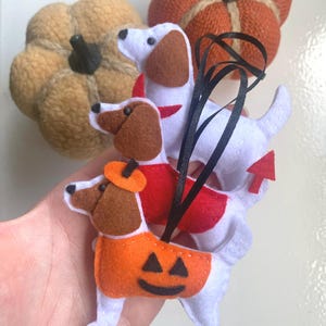 Könnte beinhalten: Drei Filz-Hundeornamente, gestapelt. Die Hunde haben weiße Körper und braune Ohren. Der oberste Hund hat ein rotes Halsband, der mittlere Hund eine rote Weste und der untere Hund ein orangefarbenes Kürbiskostüm. Zwei dekorative Kürbisse im Hintergrund.