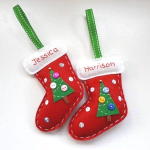 Könnte beinhalten: Zwei rote Weihnachtsstrumpf-Ornamente mit weißem Besatz und grünen Bändern zum Aufhängen. Jeder Strumpf hat einen Weihnachtsbaum aus Filz mit Knöpfen und die Namen "Jessica" und "Harrison" auf dem weißen Bund.