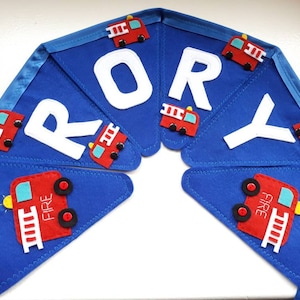 May include: A blue felt banner with the name "RORY" in white felt letters. Each letter is decorated with a red felt fire truck with black wheels and yellow ladders.