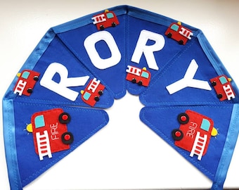 May include: A blue felt banner with the name "RORY" in white felt letters. Each letter is decorated with a red felt fire truck with black wheels and yellow ladders.