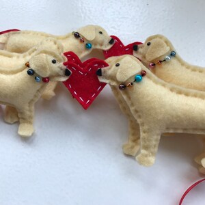 May include: Four felt dog ornaments in cream with red beaded collars, surrounding a red felt heart. The dogs are arranged in a circle around the heart.