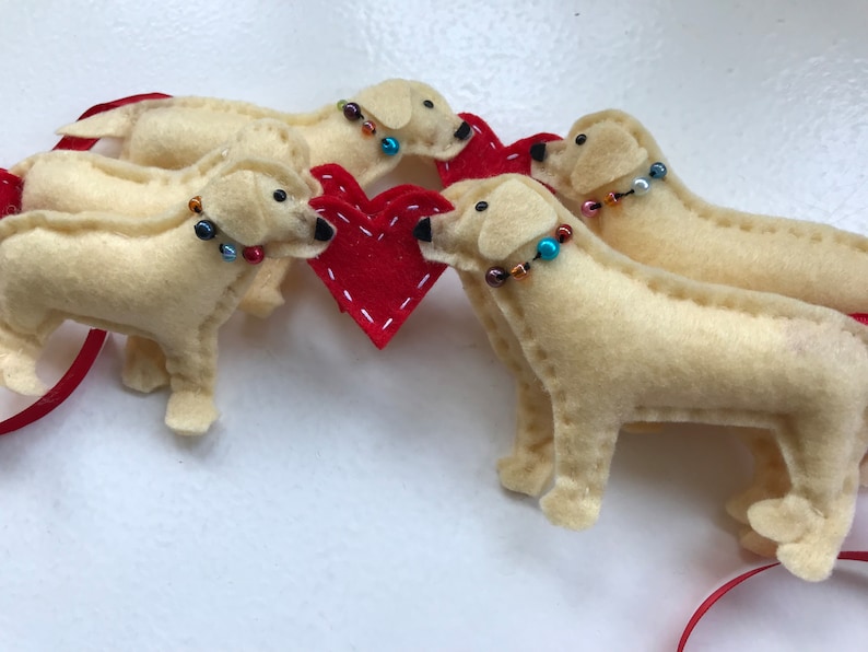 May include: Four felt dog ornaments in cream with red beaded collars, surrounding a red felt heart. The dogs are arranged in a circle around the heart.