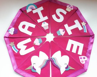 Könnte beinhalten: Ein pinkfarbenes Stoffbanner mit weißen Buchstaben, die "I SHE ME" buchstabieren. Das Banner ist mit Filz-Cupcakes, Regenbogen, Sternen und Einhornköpfen verziert. Das Banner ist mit einem pinkfarbenen Satinband eingefasst.